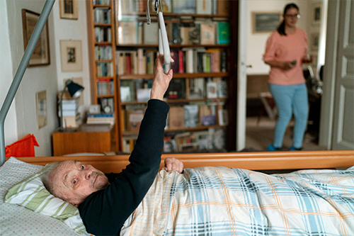 Patient liegt im Bett und hält sich an der Aufsteh-Hilfe fest. Die Pflegerin steht in der Tür.
